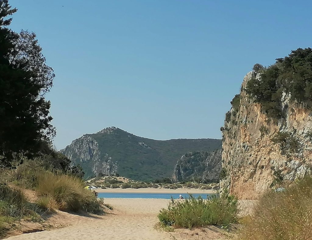 Sandy path between rocks and low vegetation leading to Voidokilia Beach